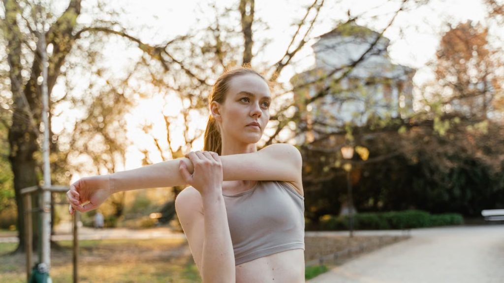 A young woman in activewear stretching her arm outdoors in a park setting during spring.